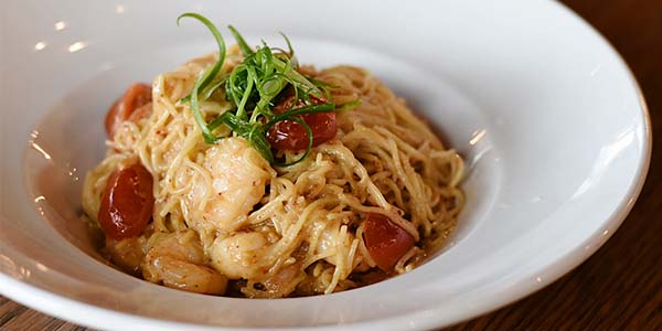 A bowl of Tom Yum Pasta: spaghetti in a Tom Yum inspired sauce with red cherry tomatoes and shrimp, topped with bright green curls of green onion.